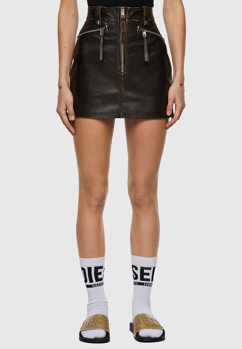 Mini jupe en cuir noir avec une taille haute, dotée de poches à double zip et de détails zippés. Associée à des chaussettes blanches et des sandales à glissière.