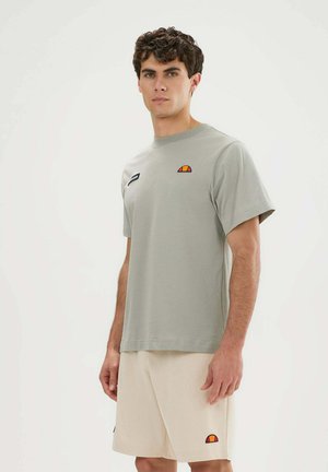 Junger Mann steht und trägt ein hellgraues kurzärmliges T-Shirt und beige Shorts, beide mit kleinen, bunten Logos auf der Vorderseite.