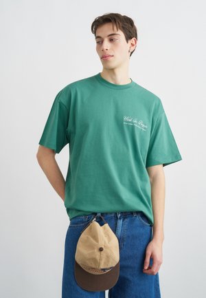 Giovane uomo con indosso una T-shirt verde con scritte e jeans blu che tiene in mano un cappellino da baseball beige e marrone su sfondo bianco.