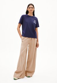 Frau steht mit Händen in den Taschen, trägt ein marineblaues T-Shirt mit weißem Text und weite beige Hosen vor einem weißen Hintergrund.