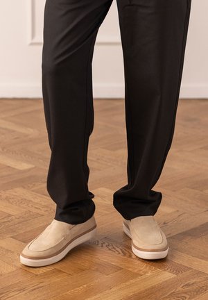 Scarpe slip-on in suede beige abbinate a pantaloni neri ampi su un pavimento in parquet di legno in un ambiente interno.