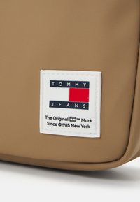 Bolsa marrón con un parche rectangular blanco con el texto "TOMMY JEANS", acentos en rojo y azul, y "The Original Mark" en negro debajo.