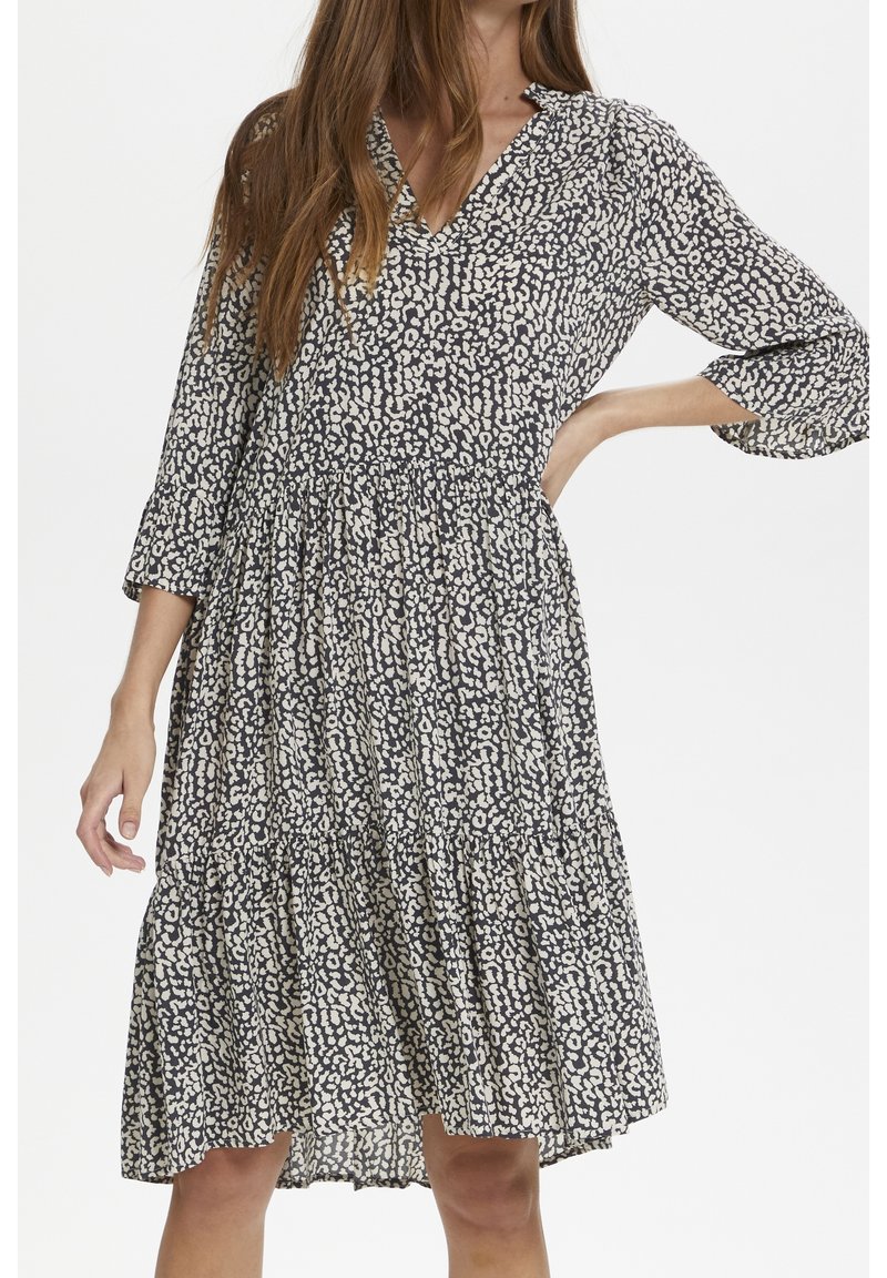 Femme portant une robe noire et blanche à pois, longueur genou, manches 3/4, encolure en V et taille froncée, posant avec une main sur la hanche.
