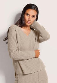 Frau mit langen, dunklen Haaren, die einen beigen, gerippten V-Ausschnitt-Pullover und einen Rock trägt, posiert mit einer Hand hinter ihrem Kopf.