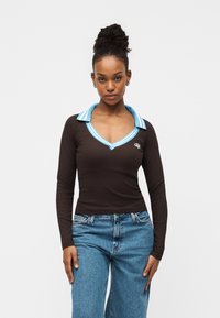 Καφέ μακρυμάνικη μπλούζα με μπλε ριγέ γιακά και λεπτομέρεια λογότυπου. Διαθέτει σχέδιο με V-λαιμόκοψη και συνδυάζεται με ανοιχτό μπλε τζιν παντελόνι.