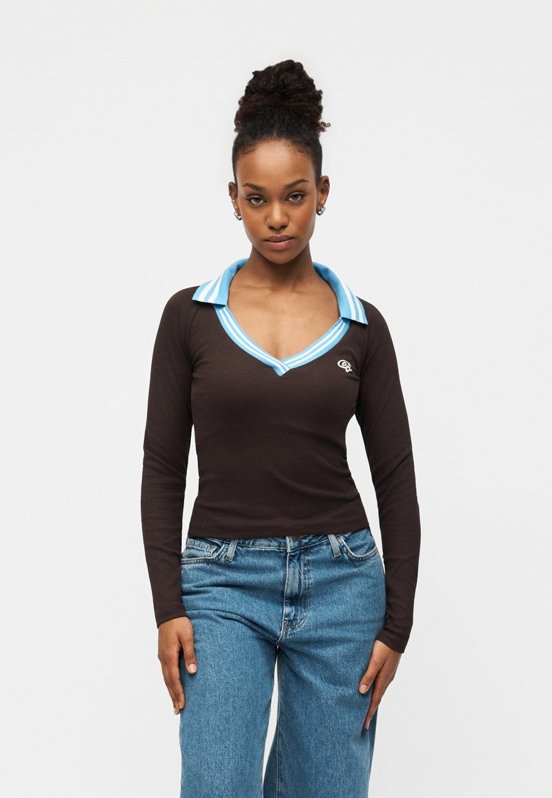 Καφέ μακρυμάνικη μπλούζα με μπλε ριγέ γιακά και λεπτομέρεια λογότυπου. Διαθέτει σχέδιο με V-λαιμόκοψη και συνδυάζεται με ανοιχτό μπλε τζιν παντελόνι.