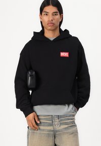 Sort hættetrøje med en frontlomme og rødt "DIESEL" logo. Båret over en grå t-shirt; parret med lyse denim jeans. Inkluderer en lille sort taske.