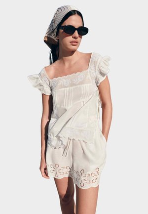 Kvinde iført hvid broderet bluse og matchende shorts med blomsterudskæringer, mørke solbriller og tørklæde med striber, vendt let mod venstre.