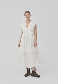 Hvit maxikjole med dyp V-hals, samlet midje og korte ermer. Stoffet er lett og semi-transparent, kombinert med brune ankelstøvler.
