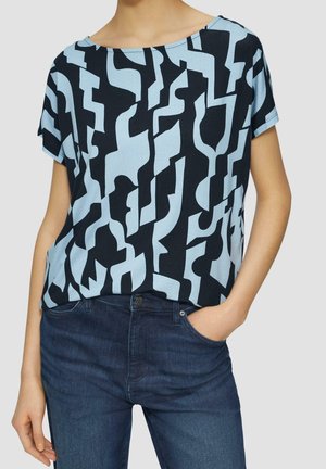 Kobieta nosząca luźną bluzkę z krótkim rękawem z abstrakcyjnym jasnoniebieskim i czarnym geometrycznym wzorem, w zestawieniu z ciemnoniebieskimi jeansami, ręka w kieszeni.