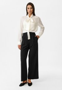 Witte blouse met lange mouwen, een gestrikte kraag en verticale strepen, gecombineerd met hoge, zwarte wijde broek en zwarte instappers.