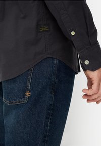 Dunkelgraues Langarmshirt mit geknöpften Manschetten, kombiniert mit blauen Denim-Jeans, die einen kleinen orangen Akzent auf der Gesäßtasche haben.