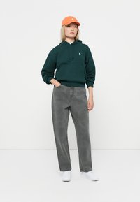 Hoodie en coton vert foncé avec poche kangourou, jean gris décontracté, et casquette orange, associé à des baskets blanches sur un fond uni.