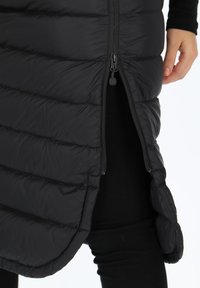 Black padded vest with a zippered front, curved hem, and quilted texture. Features a side slit for ease of movement.