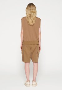 Bruine mouwloze top met een geribbelde zoom, gecombineerd met bruin cargoshorts tot de knie met zijzakken. Effen witte schoenen maken de outfit compleet.