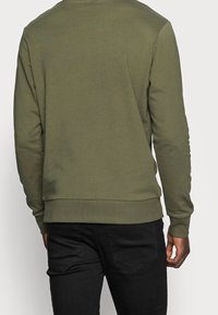 Man i olivgrön långärmad sweatshirt och svarta jeans, sedd bakifrån mot en enkel ljusbakgrund.