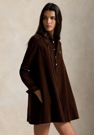 Kvinde iført en brun langærmet, knap-til kjole med krave, stående med den ene hånd i lommen, kigger til siden mod en beige baggrund.