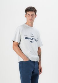 Grauer Baumwoll-T-Shirt mit kurzen Ärmeln, versehen mit einem dunkelblauen "ATHLETICS"-Druck und kleinen Textdetails, kombiniert mit dunkelblauen Jeans.