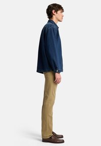 Giacca di jeans blu scuro con vestibilità rilassata, dotata di maniche lunghe e polsini con bottone, abbinata a pantaloni khaki e mocassini marroni.