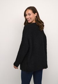 Schwarzer gestrickter Pullover mit strukturierter Musterung, lässiger Schnitt und geripptem Saum. Getragen mit blauen Jeans, von hinten gezeigt.