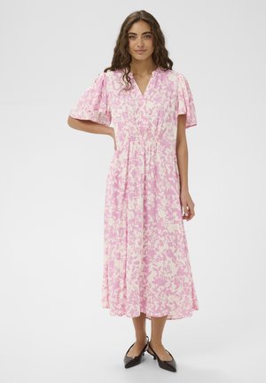 Kobieta o długich kręconych włosach, ubrana w różowo-białą kwiecistą sukienkę midi i czarne buty na niskim obcasie, stojąca z jedną ręką na biodrze.