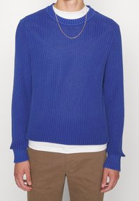 Pull en maille bleu avec une texture côtelée, col rond et manches longues. Porté par-dessus une chemise blanche, avec une chaîne dorée discrète.