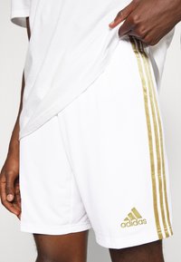 Vita atletiska shorts med gyllene sidsremsor och en broderad gyllene Adidas-logotyp. Har ett andningsbart material och en bekväm passform.