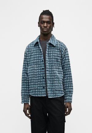 Νέος άνδρας με πλεξούδες, φορώντας μπλε καρό σακάκι πάνω από σκουρόχρωμο πουκάμισο και μαύρο παντελόνι, στέκεται μπροστά σε ένα απλό λευκό φόντο.