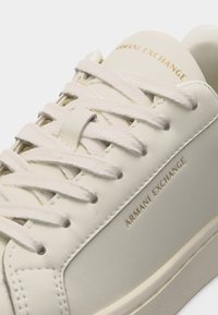 Κοντινή λήψη λευκού αθλητικού παπουτσιού Armani Exchange που δείχνει τα κορδόνια και το χρυσό λογότυπο στην γλώσσα και στην πλαϊνή επιφάνεια.