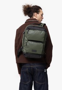 Grön nylonryggsäck med två frampocket, svarta accenter, justerbara remmar och en texturerad botten, bärs på en persons rygg.
