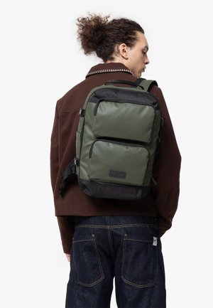 Green nylon backpack with two front pockets, black accents, adjustable straps, and a textured bottom, worn on a person’s back.