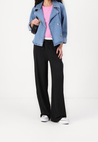 Chaqueta de denim de doble botonadura, camiseta rosa, pantalones anchos de rayas negras, y zapatillas blancas. Sostiene un bolso negro. Fondo plano.