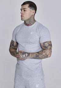 Camiseta gris ajustada hecha de tela suave, con un pequeño logo blanco en el lado izquierdo del pecho, con una textura lisa y un cuello redondo.