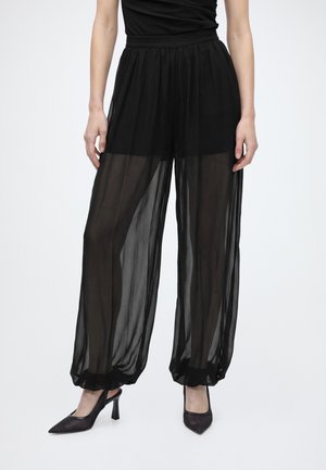 Pantalon large noir transparent avec chevilles froncées porté avec des chaussures à talons noirs et un top noir ajusté, visible de la taille aux pieds.