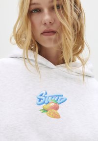Giovane donna con capelli biondi indossa un felpa grigia chiara con il testo blu "STWD" e una grafica di mango sul petto, su sfondo bianco.