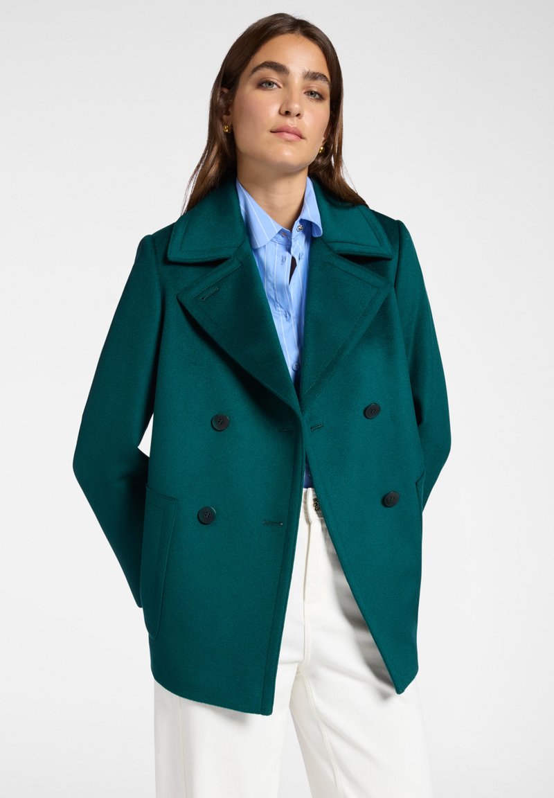 Donna che indossa un cappotto doppiopetto color teal, una camicia a righe blu e pantaloni bianchi, che si trova davanti a uno sfondo chiaro e semplice.