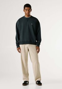 Felpa blu scuro con polsini a coste, dotata di un piccolo logo sul petto, abbinata a pantaloni larghi con coste beige e sneakers nere.