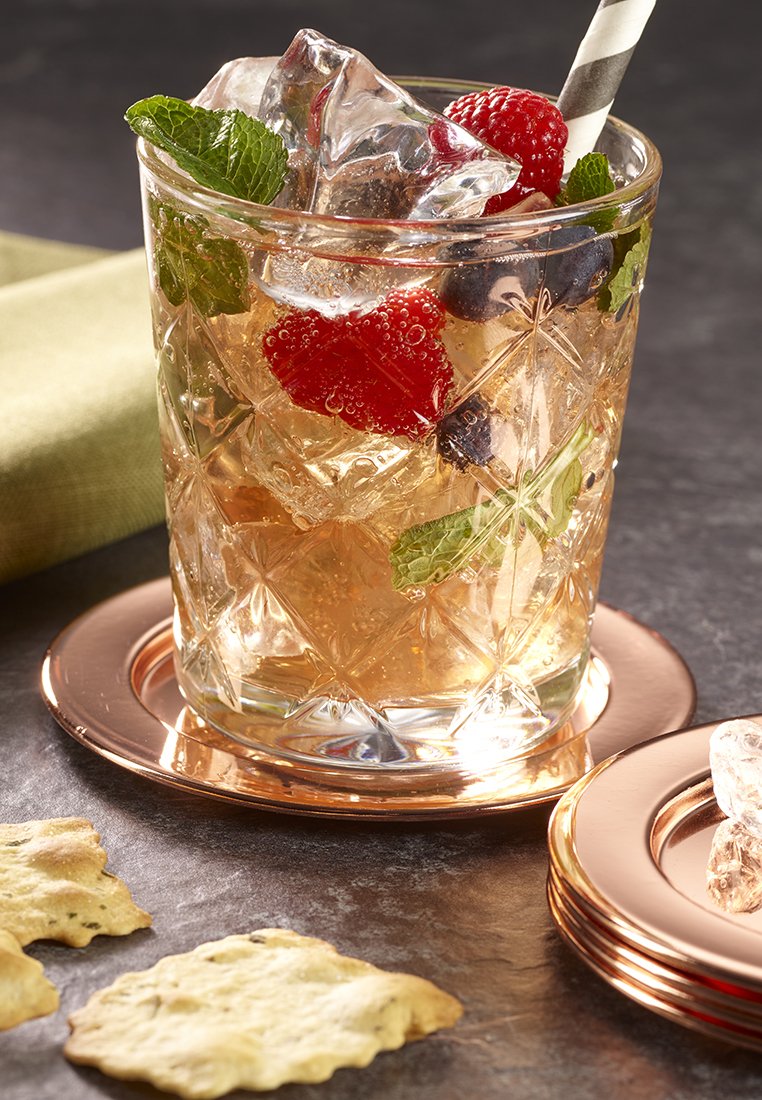 Glas mit einem eisgekühlten Beerendrink, garniert mit Minzblättern und einem gestreiften Strohhalm, auf einem bronzenen Untersetzer, neben Kräcker auf einer marmorierten Oberfläche.