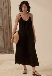 Robe maxi noire sans manches en tissu doux et texturé avec un décolleté plongeant. Assortie avec un sac en tricot beige et des tongs noires.