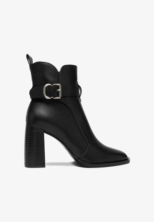Bota de cuero negro hasta el tobillo con un tacón alto y ancho, puntera puntiaguda y una correa decorativa con hebilla plateada alrededor del tobillo.