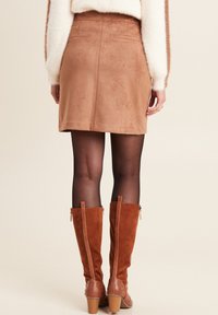 Frau in einem beigen Wildleder-Minirock, schwarzen durchsichtigen Strumpfhosen, braunen kniehohen Stiefeln mit Blockabsätzen und einem weißen flauschigen Pullover, die von hinten steht.