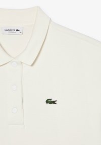 Polo shirt color crema con finitura testurizzata, caratterizzato da un colletto con tre bottoni e un piccolo logo di coccodrillo ricamato in verde.