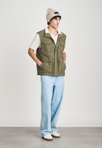 Ελαιόχρωμο μαλακό γιλέκο με πολλά μπροστινά τσεπάκια, φορεμένο πάνω από ένα λευκό πουκάμισο και ανοιχτό μπλε τζιν, συνδυασμένο με λευκά αθλητικά παπούτσια και μπεζ σκούφο.