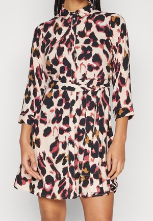 Femme portant une robe ceinturée à boutons sur le devant, avec des manches trois-quarts, présentant un motif abstrait noir, rose et marron sur fond beige.