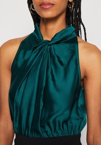 Tyrkysový saténový top na halter s překrouceným výstřihem a nařaseným pasem. Hladká textura a bezrukávový design, který zvýrazňuje ramena.