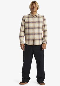 Camisa de franela de cuadros beige y púrpura con botones en la parte delantera, un bolsillo en el pecho y pantalones oscuros de corte relajado. Zapatos marrones.