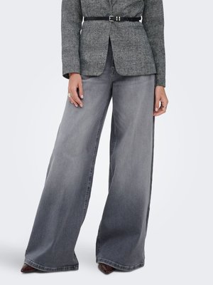 Mujer con vaqueros gris desgastados de pierna ancha, blazer gris hecho a medida con cinturón negro y zapatos marrones puntiagudos, de pie frente a un fondo claro.