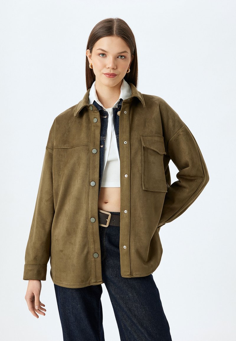 Giacca in finta suede verde oliva, morbida, con colletto, bottoni a scatto frontali, tasca sul petto e vestibilità comoda. Indossata sopra un top bianco e jeans scuri.