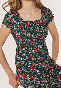 Sukienka w kwiatowy wzór na zielonym tle z małymi czerwonymi, różowymi, pomarańczowymi i fioletowymi kwiatami. De kolt ozdobiony marszczeniem i bufiaste rękawy.