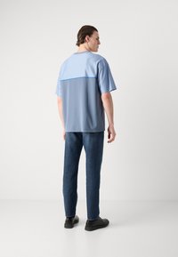 Short-sleeve, light blue t-shirt with textured upper panel and solid lower section. Worn with dark blue jeans and black shoes.